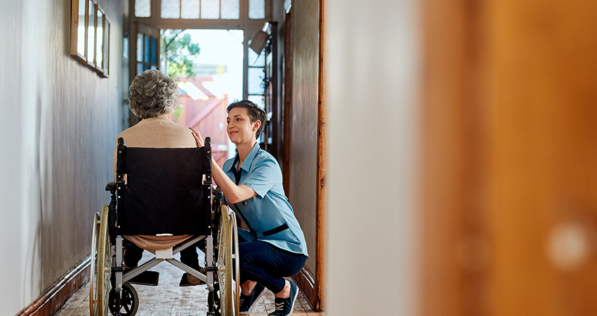 A caregiver kneels beside an elderly person in a wheelchair, offering support in a sunlit hallway of what appears to be a care facility or home, embodying the dedication essential for effective care gap closure and patient assessment.