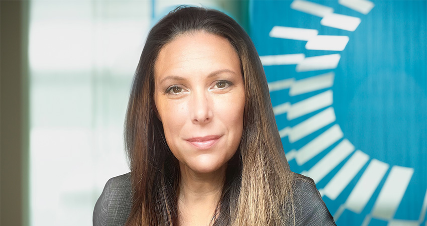 A woman with long brown hair and a slight smile poses in front of a blue background with white geometric patterns, subtly evoking the precision and clarity found in AHRQ quality indicators.