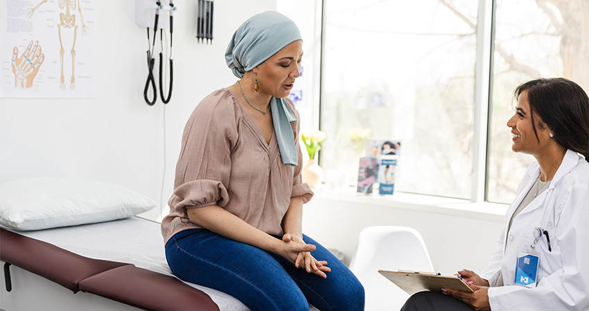 A woman wearing a headscarf sits on an exam table, speaking with a doctor who holds a clipboard in a bright medical office, as they discuss the importance of considering SDOH in cancer care during her treatment planning.