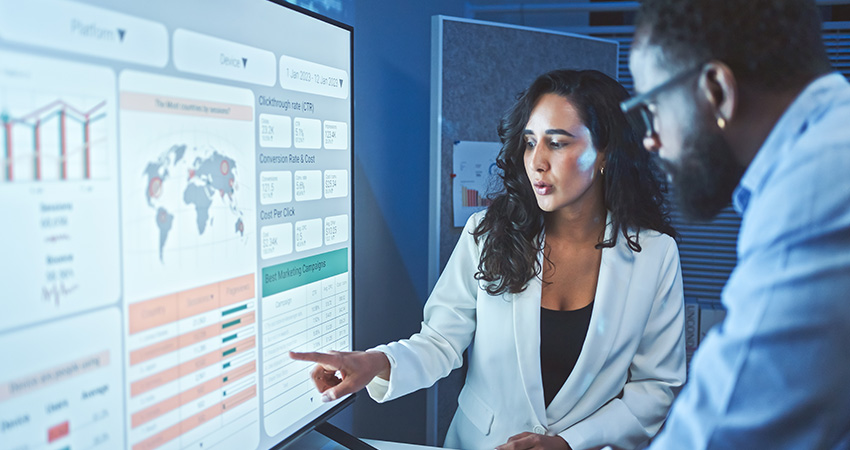 Two people analyze data on a digital screen displaying charts, graphs, a world map, and statistics related to clinical trial recruitment in an office setting. One person points at the screen while discussing the information with the other.