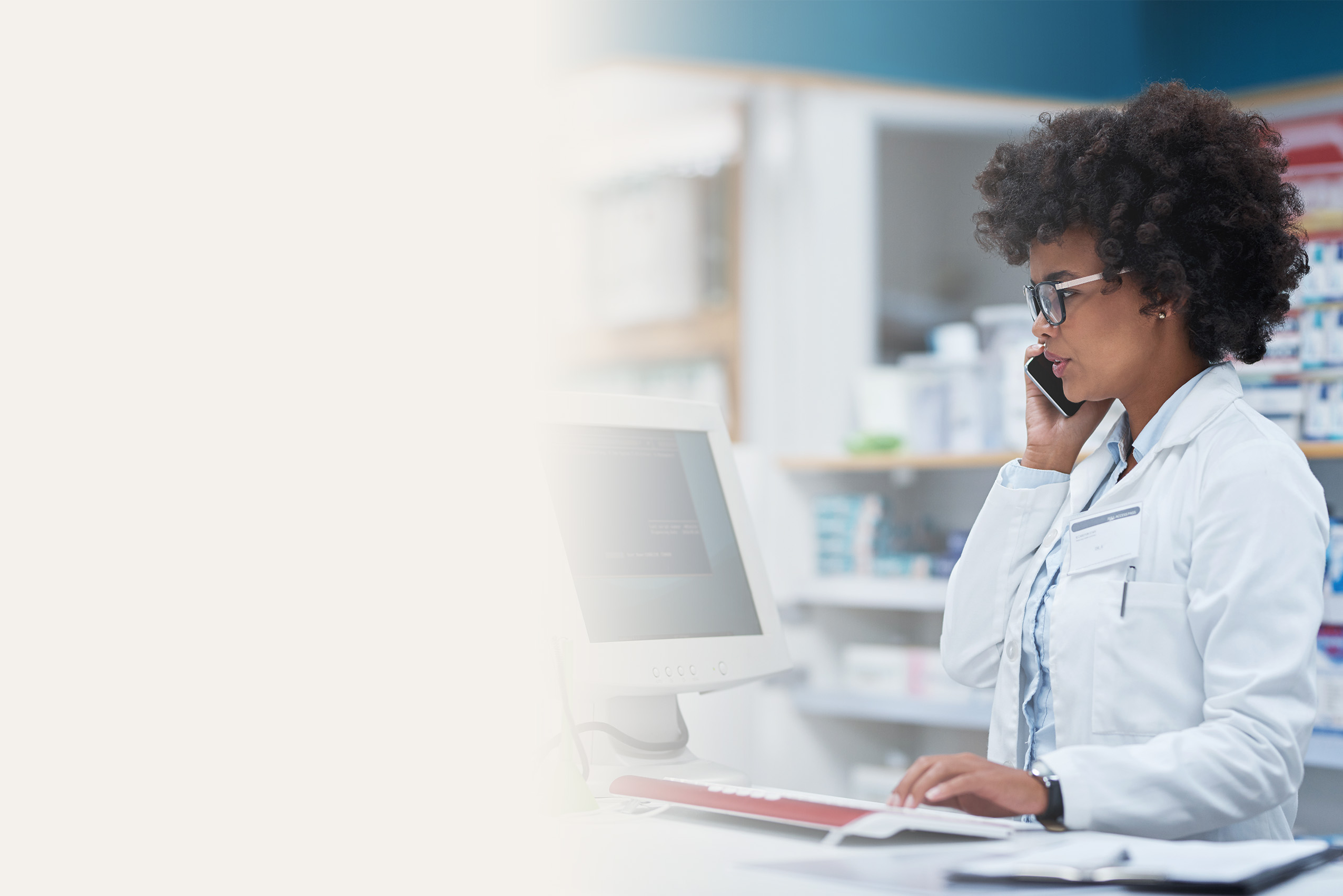 A pharmacist in a white coat talks on the phone while using a computer equipped with pharmacy management software at a pharmacy counter, with medicine shelves visible in the background.