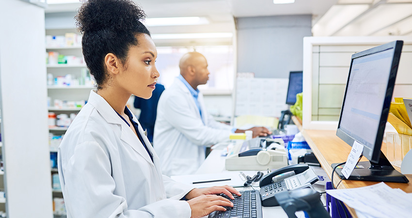 A woman in a white lab coat types on a keyboard in a pharmacy, with shelves of medicines and another person working in the background.
