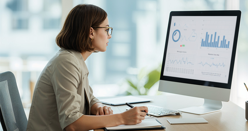 A woman sits at a desk, taking notes while looking at real-world data displayed as graphs and charts on a computer monitor in a bright office setting.