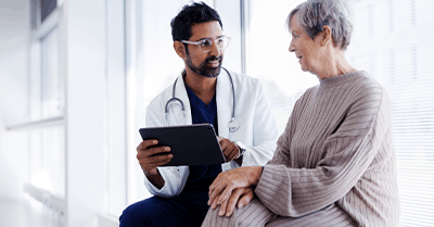A doctor holding a tablet speaks with an older woman in a light-filled medical office. The woman is sitting and listening attentively, while the doctor smiles and explains something.