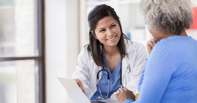 A doctor smiles and talks with a patient, holding a clipboard. The doctor wears a white coat and stethoscope, while the patient, seen from behind, has gray hair and wears a blue shirt.