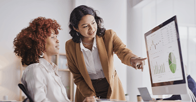 Two women in an office look at a computer screen displaying graphs and charts. One woman stands, pointing at the monitor, while the other sits and listens attentively.