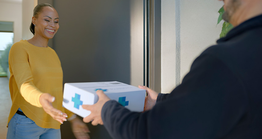 Smiling woman in a yellow shirt at her front door receiving a white delivery box with blue medical-style cross symbols from a courier.