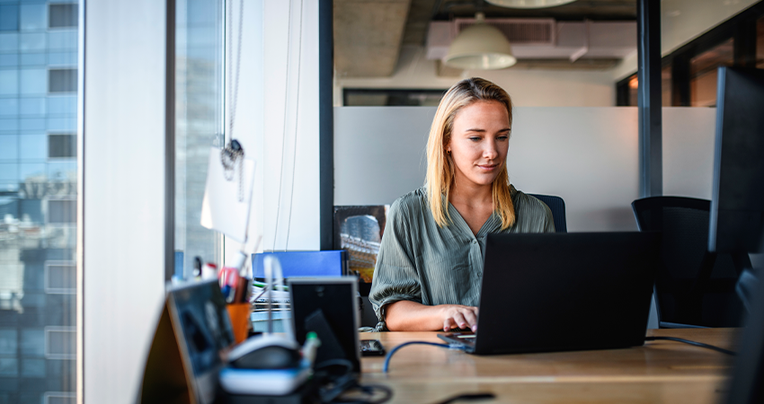 A woman with blonde hair is sitting at a desk, typing on a laptop.