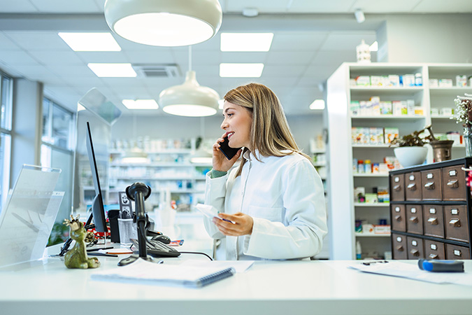 A pharmacist in a white coat talks on the phone while holding a prescription at a pharmacy counter with a computer, scanner, and shelves of medications in the background.