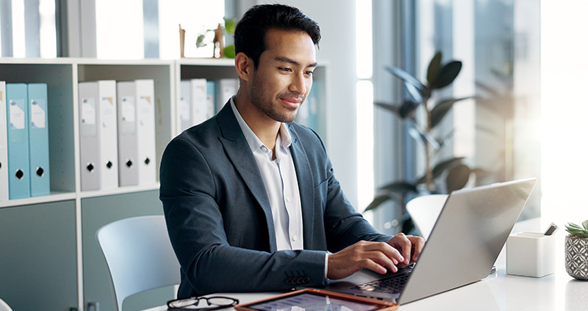 A man in a suit sits at a desk in an office, typing on a laptop. There are binders on shelves and a potted plant in the background.