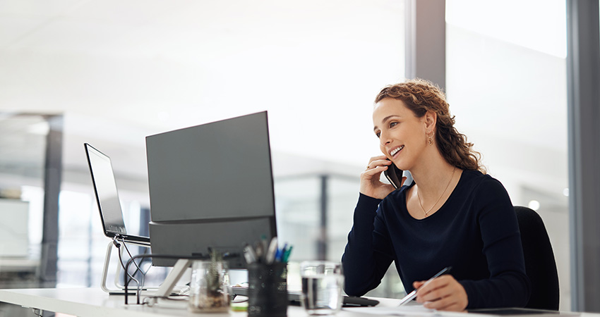 A woman sitting at a desk in an office talks on the phone while taking notes about strategies to reduce claim denials in ASCs, with a computer monitor and office supplies visible in front of her.