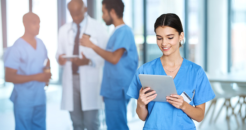 A smiling healthcare worker in blue scrubs uses a tablet, demonstrating effective nurse retention strategies, while three medical professionals talk in the background in a bright hospital setting.