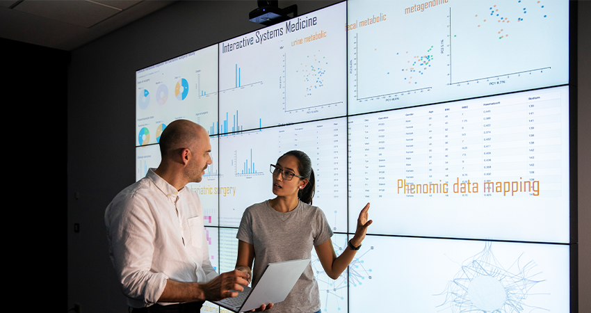Two people discuss data in front of large screens displaying charts, graphs, and the text Phenomic data mapping. One person holds a laptop while the other gestures toward the displays, illustrating how advanced analytics can enhance clinical trial patient recruitment.