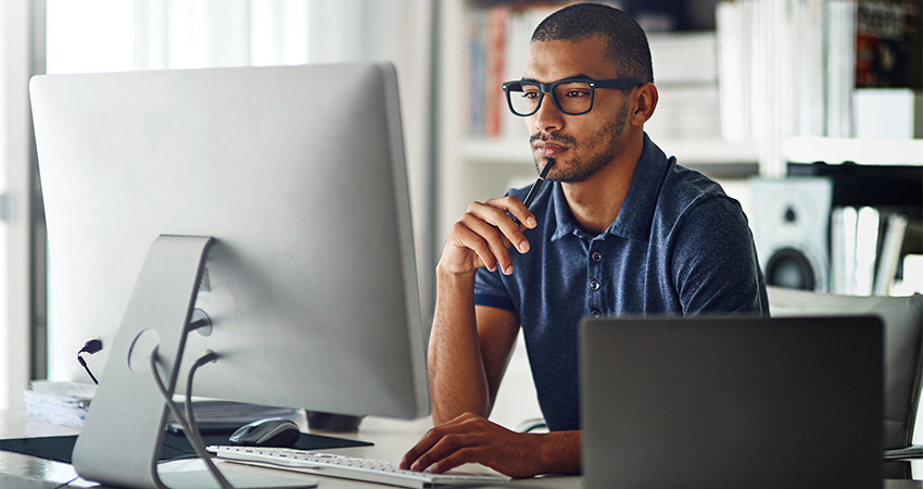 A man wearing glasses sits at a desk, looking thoughtfully at a large computer monitor with a pen resting on his chin. Surrounded by a laptop, office supplies, and open tabs displaying mobile scheduling software, he works in a modern, well-lit workspace.