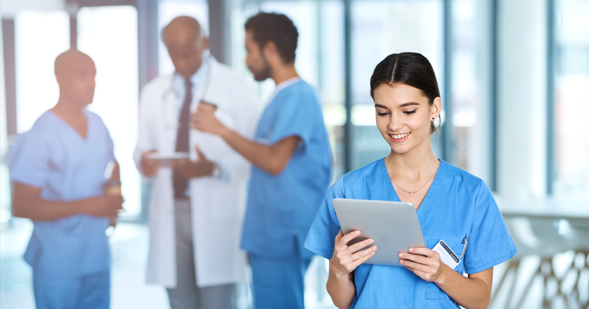 Nurse looking at tablet with doctor and nurses discussing in background