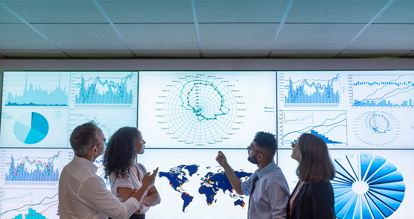 Four people stand and discuss rare disease patient data in front of large screens displaying various charts, graphs, and a world map in a modern office setting.