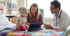 A young child with a nasal tube sits on a hospital bed, using a tablet with a woman next to her. A doctor in a white coat sits nearby, talking with them. Medical equipment is visible in the background, highlighting the dedicated care provided to rare disease patients.