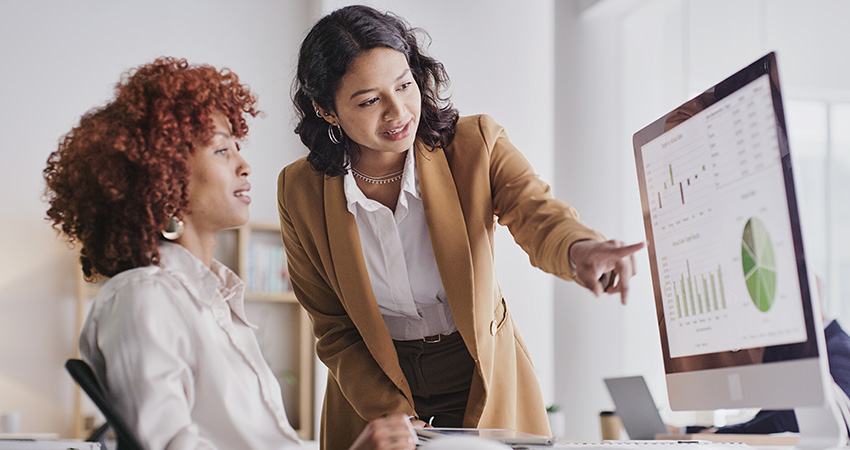 Two women in an office look at a computer screen displaying charts and graphs related to the MSSP 2027 Proposed Rule. One woman is seated, while the other stands and points at the monitor, discussing data. Bright, modern workspace environment.