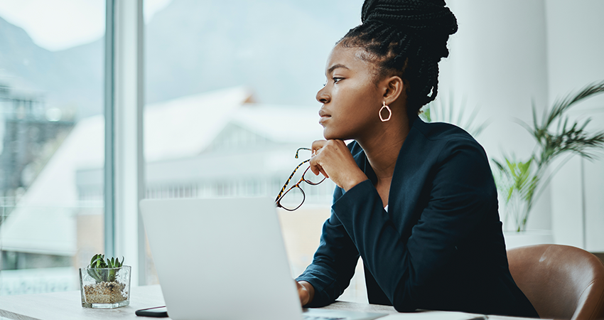 A woman sits at a desk with a laptop, holding her glasses and looking thoughtfully out a large window. With Patient Statement Delivery on the screen, she considers her next steps. There is a small plant on the desk and natural light fills the modern office space.
