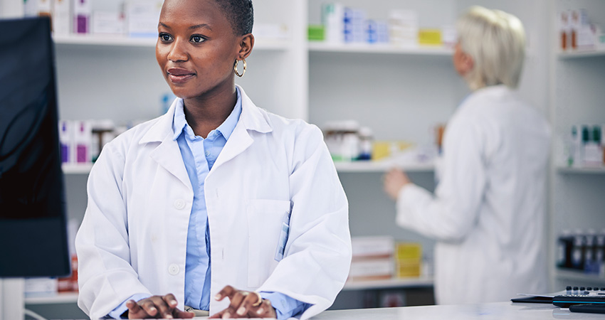 A pharmacist in a white coat works at a computer in a pharmacy, demonstrating AI readiness in specialty pharmacy, with shelves of medications behind her and another pharmacist organizing items in the background.