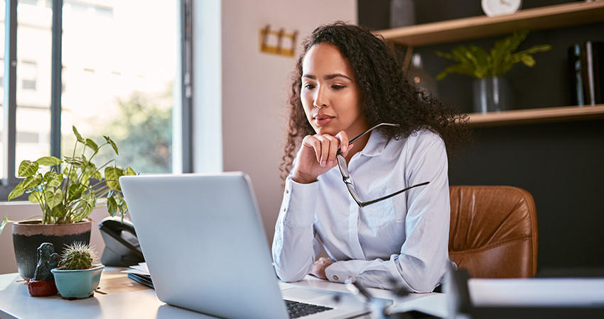 A woman with curly hair sits at a desk in an office, looking thoughtfully at her laptop. There are plants and office supplies on the desk and shelves behind her. Sunlight comes in through a nearby window.