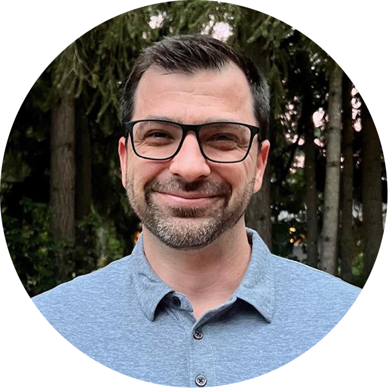 A man with short dark hair, glasses, and a beard smiles while wearing a gray collared shirt, standing outdoors in front of tall green trees, reminiscent of the natural backdrops often found in Karen Biesack’s photography.